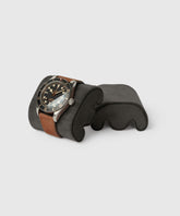 A TAWBURY Fraser Black wristwatch with a brown leather strap and black dial elegantly rests on a charcoal Fraser Watch Case Pillow, blending into the neutral backdrop. Perfect for style aficionados, it pairs beautifully with Leather Watch Travel Cases for globe-trotting adventures.